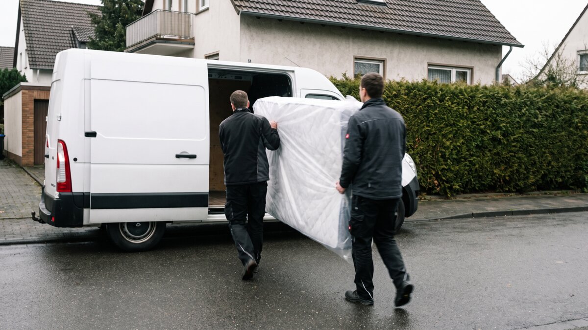 Zwei Träger bringen eine verpackte Matratze zum Transporter